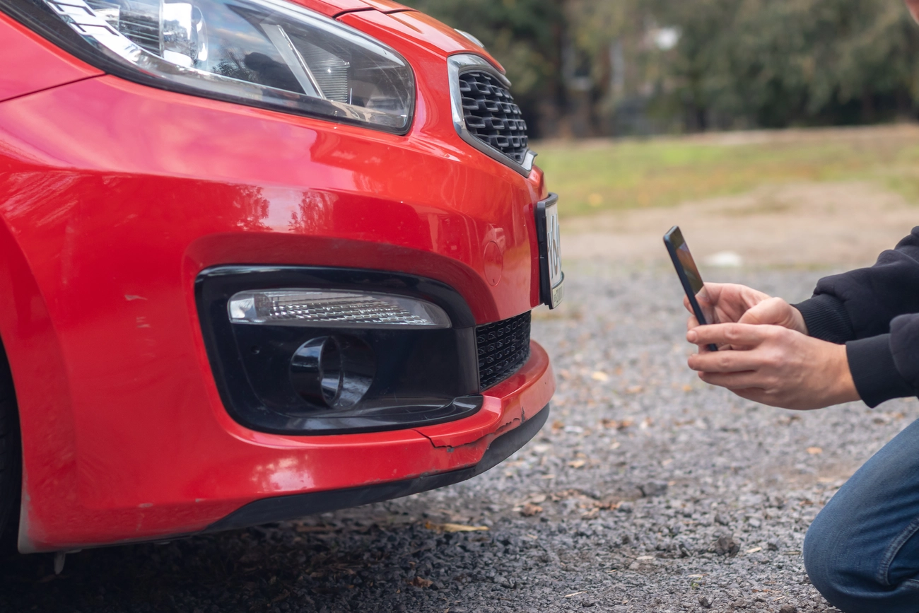 KFZ-Sachverständigenbüro - Peter Kühn - Mann fotografiert beschädigtes rotes Auto mit Smartphone. Rückansicht.